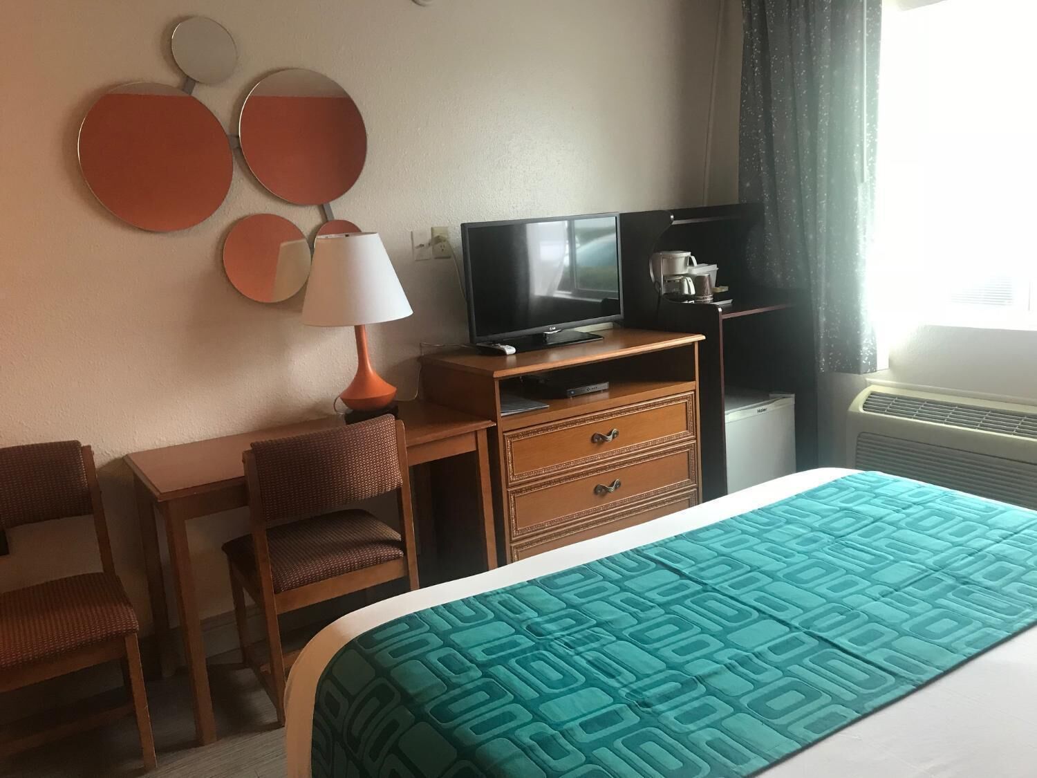 Una habitación de hotel con cama, escritorio, silla, televisor, lámpara y cafetera.