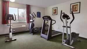 Een sportschool met loopbanden, een elliptische trainer en een stair climber. Er is een raam met gordijnen, een radiator en ingelijste foto’s aan de muur.