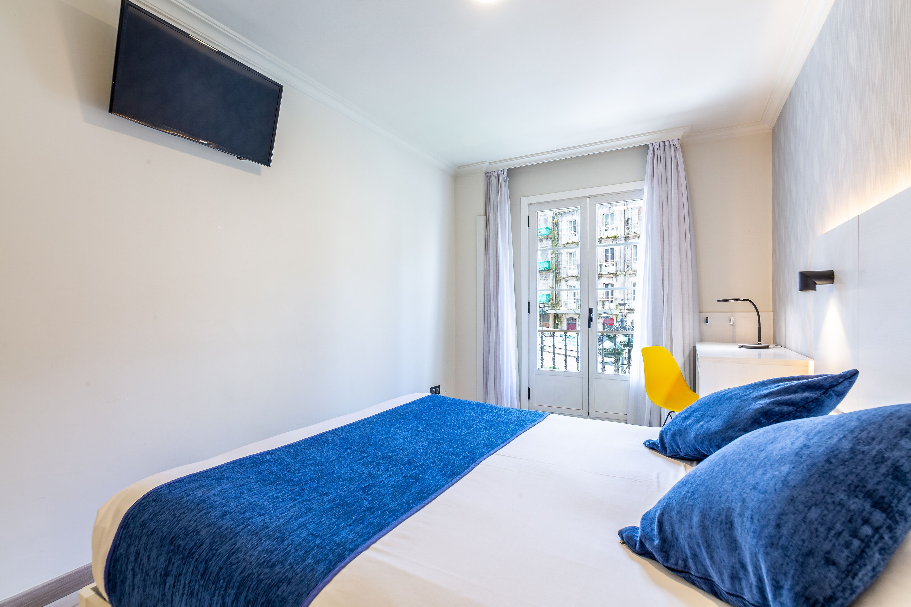 A hotel room with a bed, a flat-screen TV mounted on the wall, a window with curtains, and a view of buildings outside.