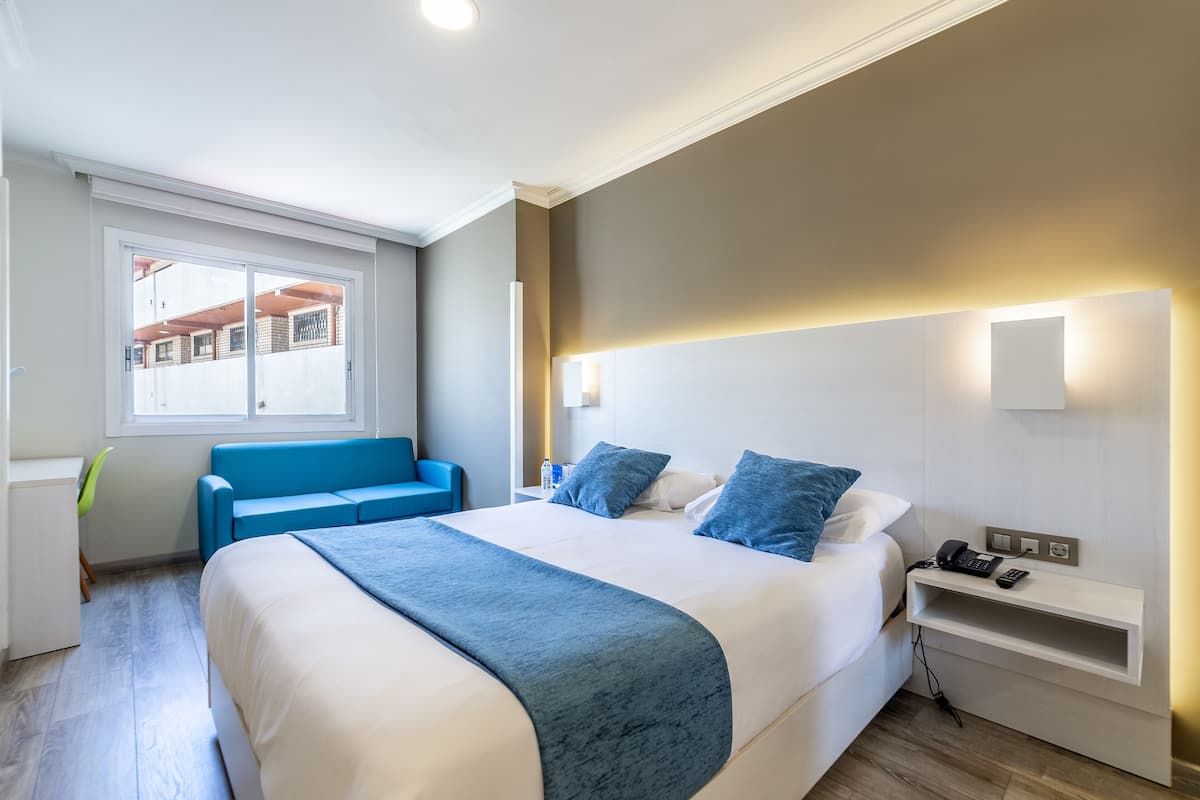 Ein modernes Hotelzimmer mit einem großen Bett, einem blauen Sofa, einem Nachttisch mit Telefon und einem Fenster mit Blick auf Gebäude.