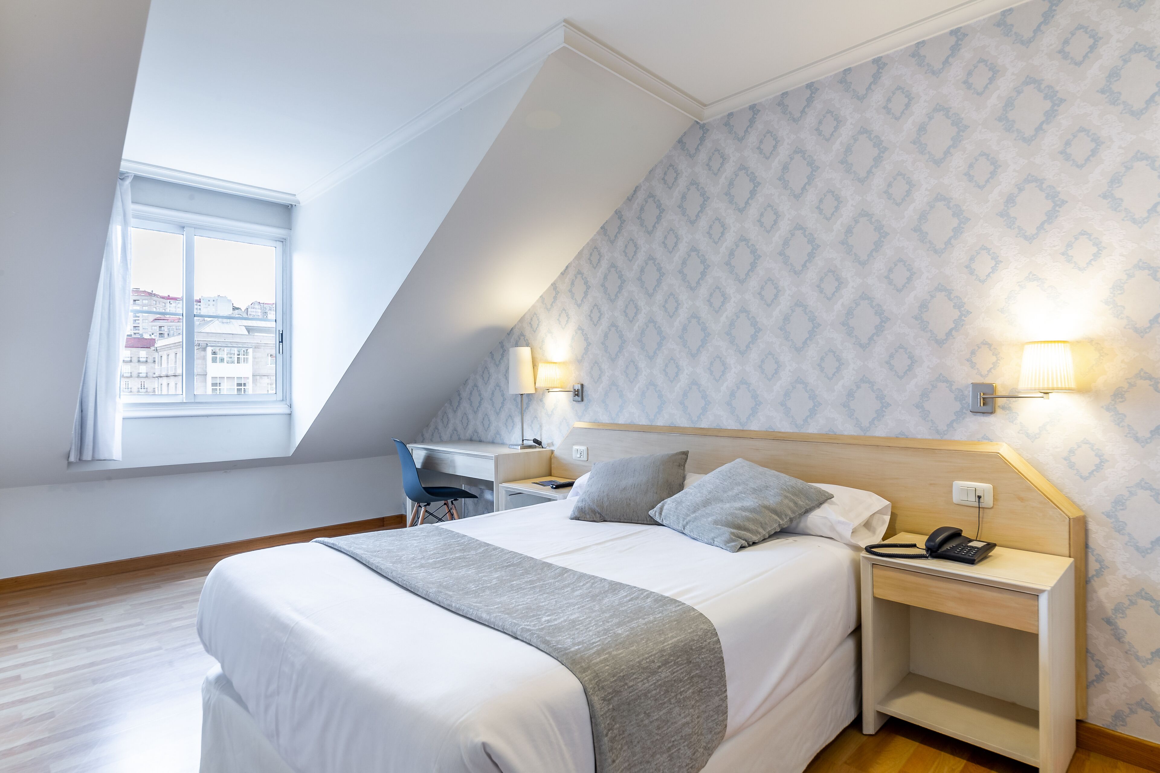 Un dormitorio con cama, escritorio y silla, una ventana con vistas, y papel tapiz estampado.