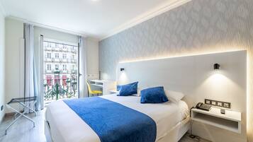 Una habitación de hotel con cama, mesita de noche, lámpara y vistas a la ciudad a través del ventanal.