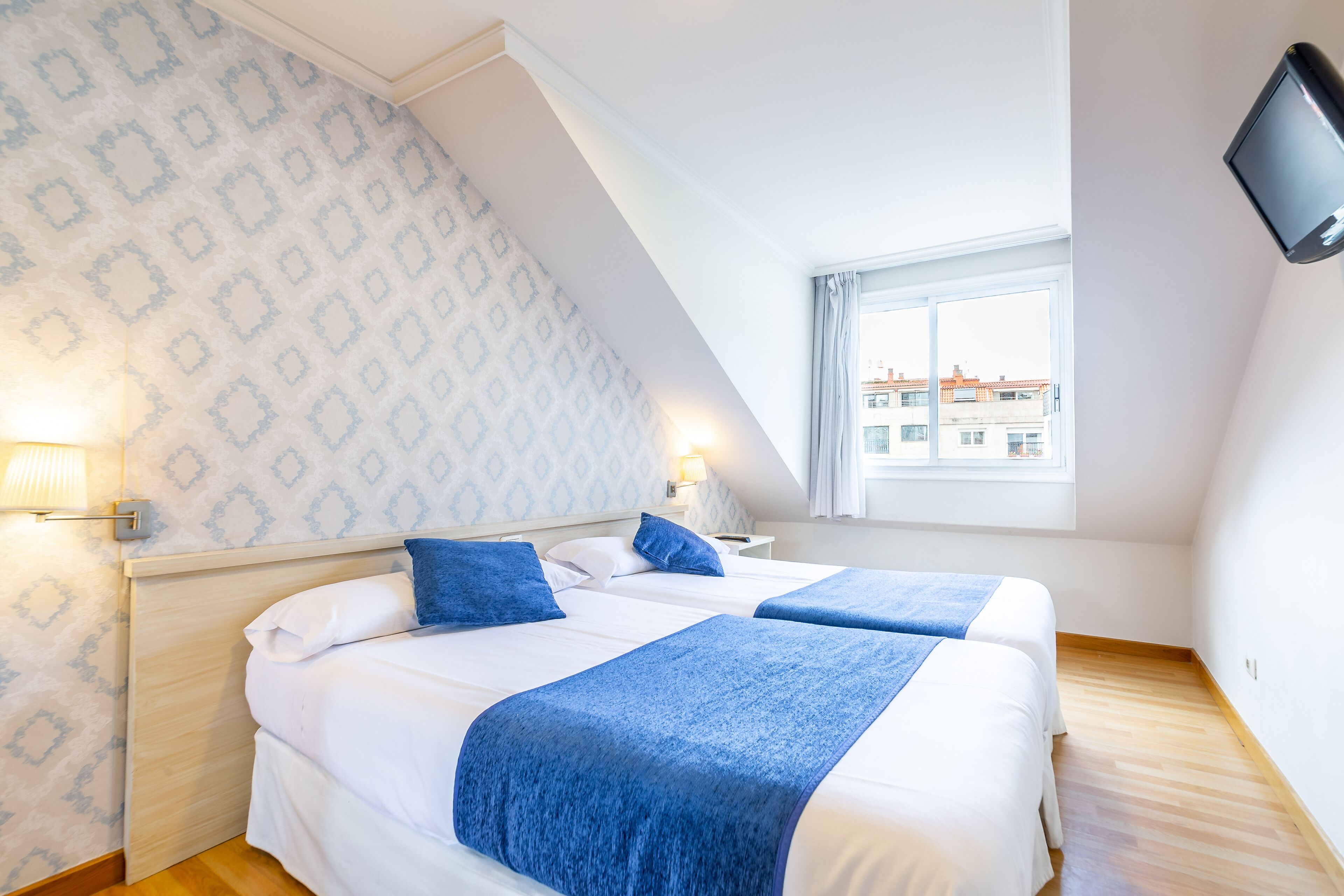 A hotel room with two beds, a TV mounted on the wall, and a window with a view of buildings.