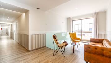 Un hall d’hôtel avec un plancher en bois, une cloison en verre, un fauteuil jaune, un canapé marron et un couloir menant à une autre pièce.