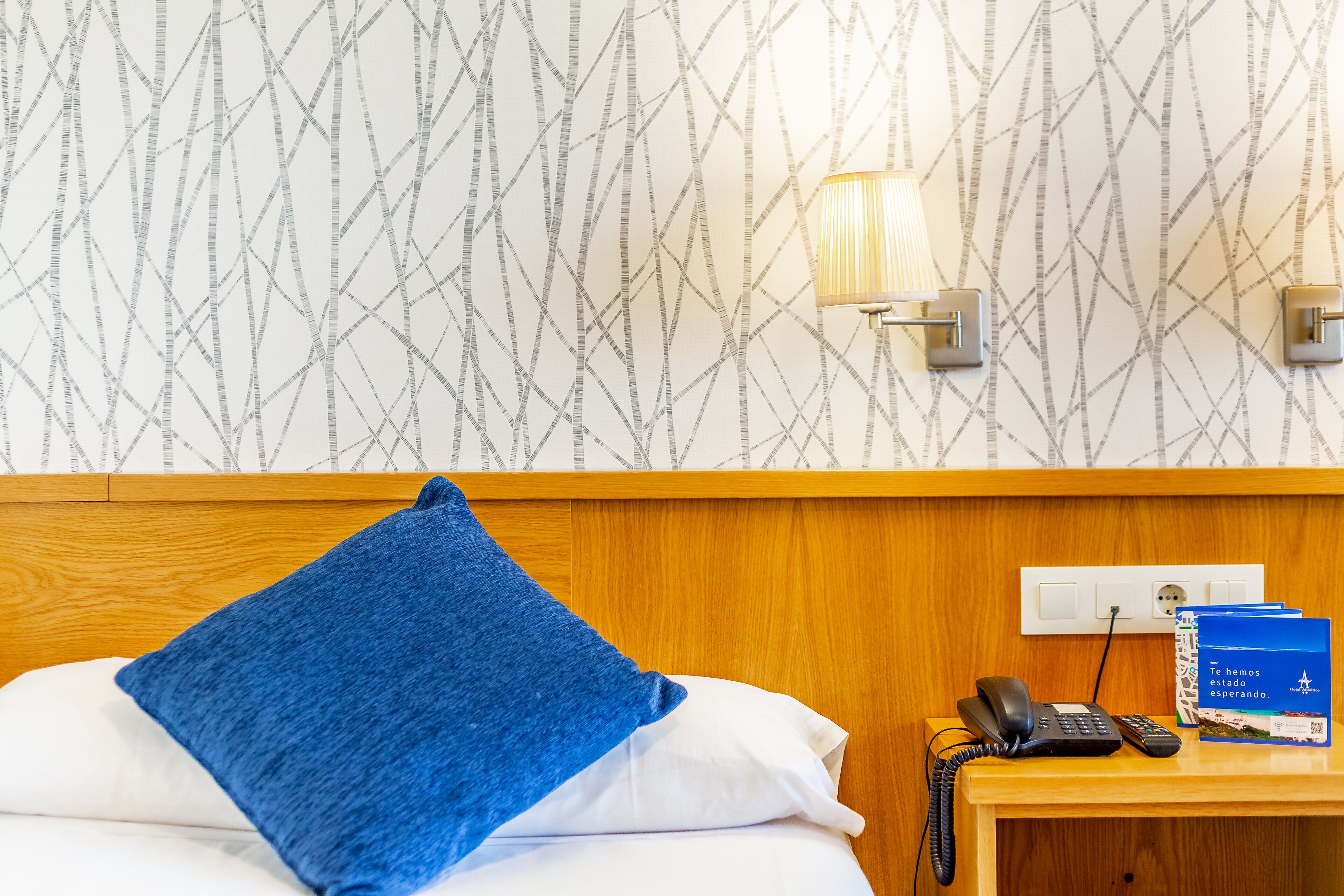 A hotel room with a wooden headboard, a blue throw pillow, a bedside table with a phone, a remote control, and a brochure.