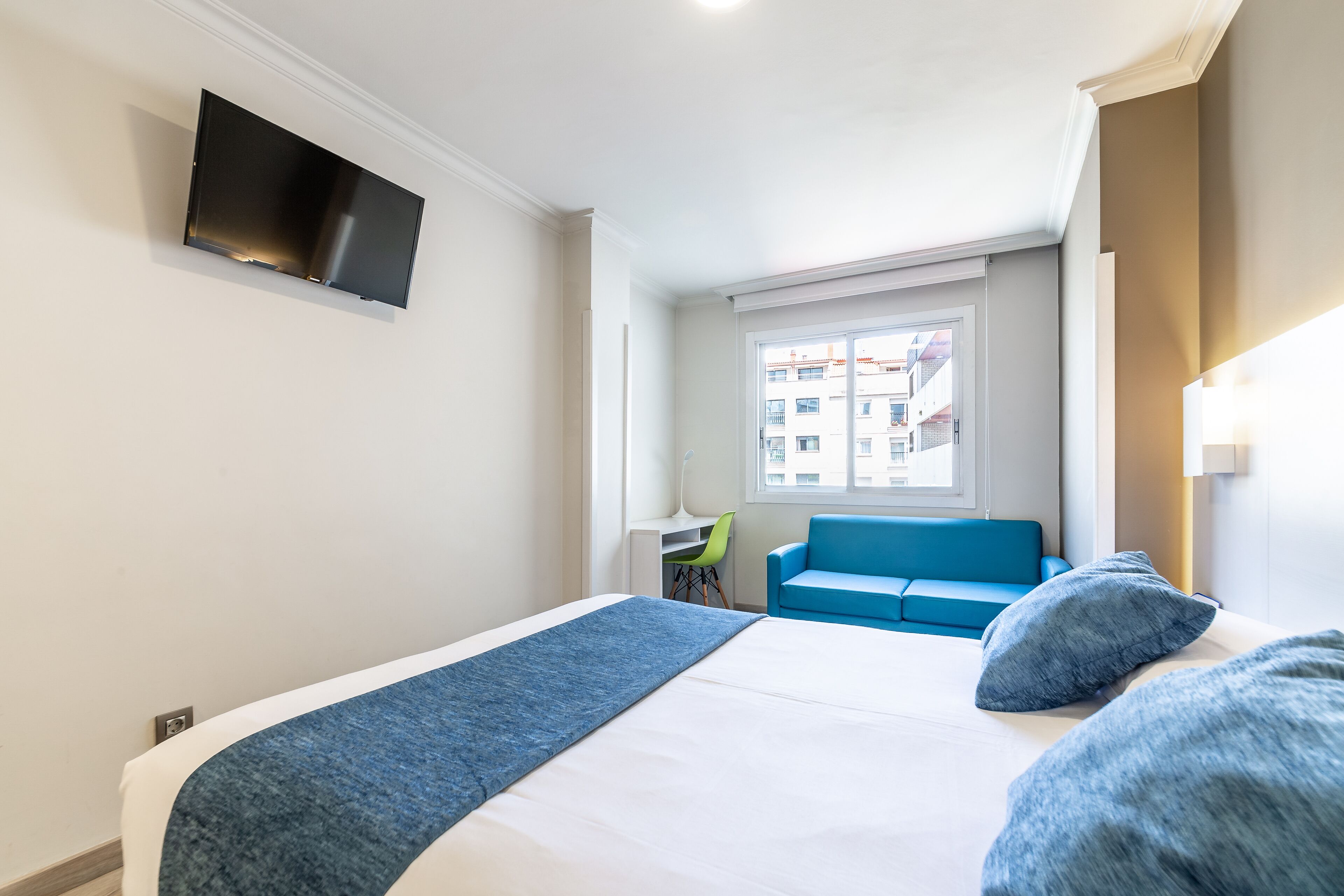 Habitación de hotel con cama, televisor montado en la pared, ventana con vistas a edificios y sofá azul.