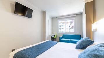 Habitación de hotel con cama, televisor montado en la pared, ventana con vistas a edificios y sofá azul.