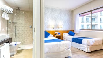 Habitación de hotel con dos camas, una ducha, una mesita con teléfono y ventana con vistas a edificios.