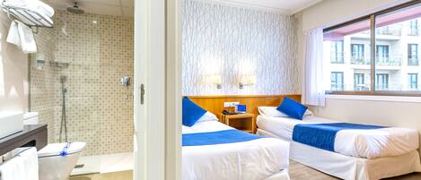 A hotel room with two beds, a shower, a small table with a phone, and a window with a view of buildings.