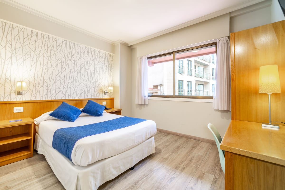 Ein Hotelzimmer mit Bett, Nachttischen, Schreibtisch, Stuhl, einem Fenster mit Vorhängen und Blick auf Gebäude.
