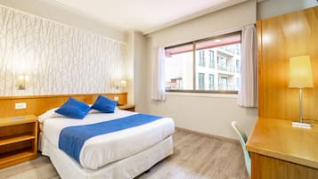 Una habitación de hotel con cama, mesitas de noche, escritorio, silla y ventana con vistas a edificios, además de una pared con diseño.