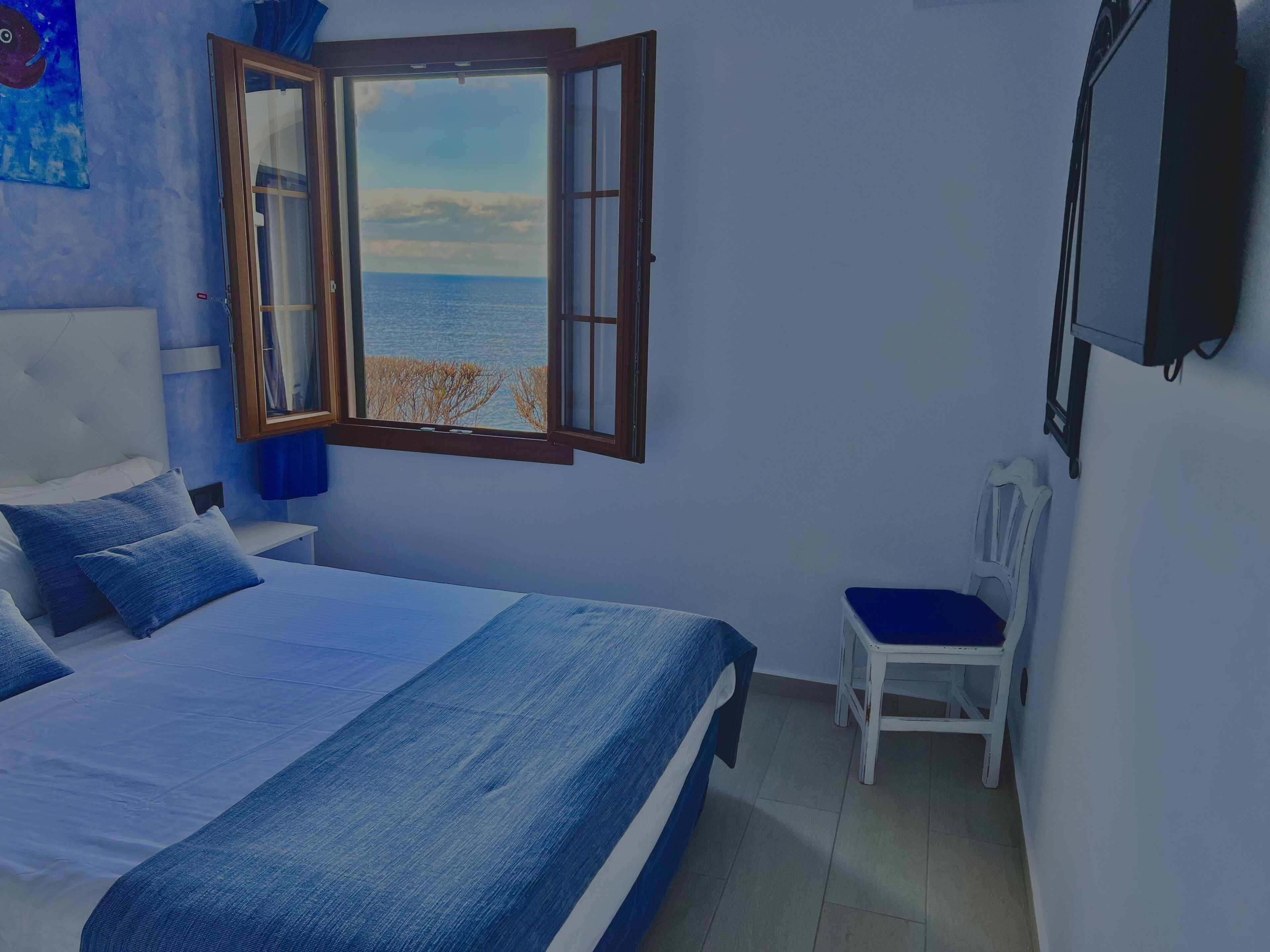 A bedroom with a bed, a chair, a TV, and a window with a view of the sea.