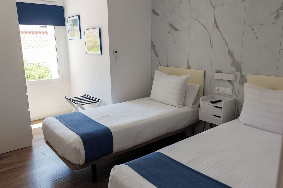 Ett hotellrum med två separata sängar, ett litet nattduksbord med en lampa, ett fönster med utsikt över grönområden och en vägg med ett mönster som liknar marmor.