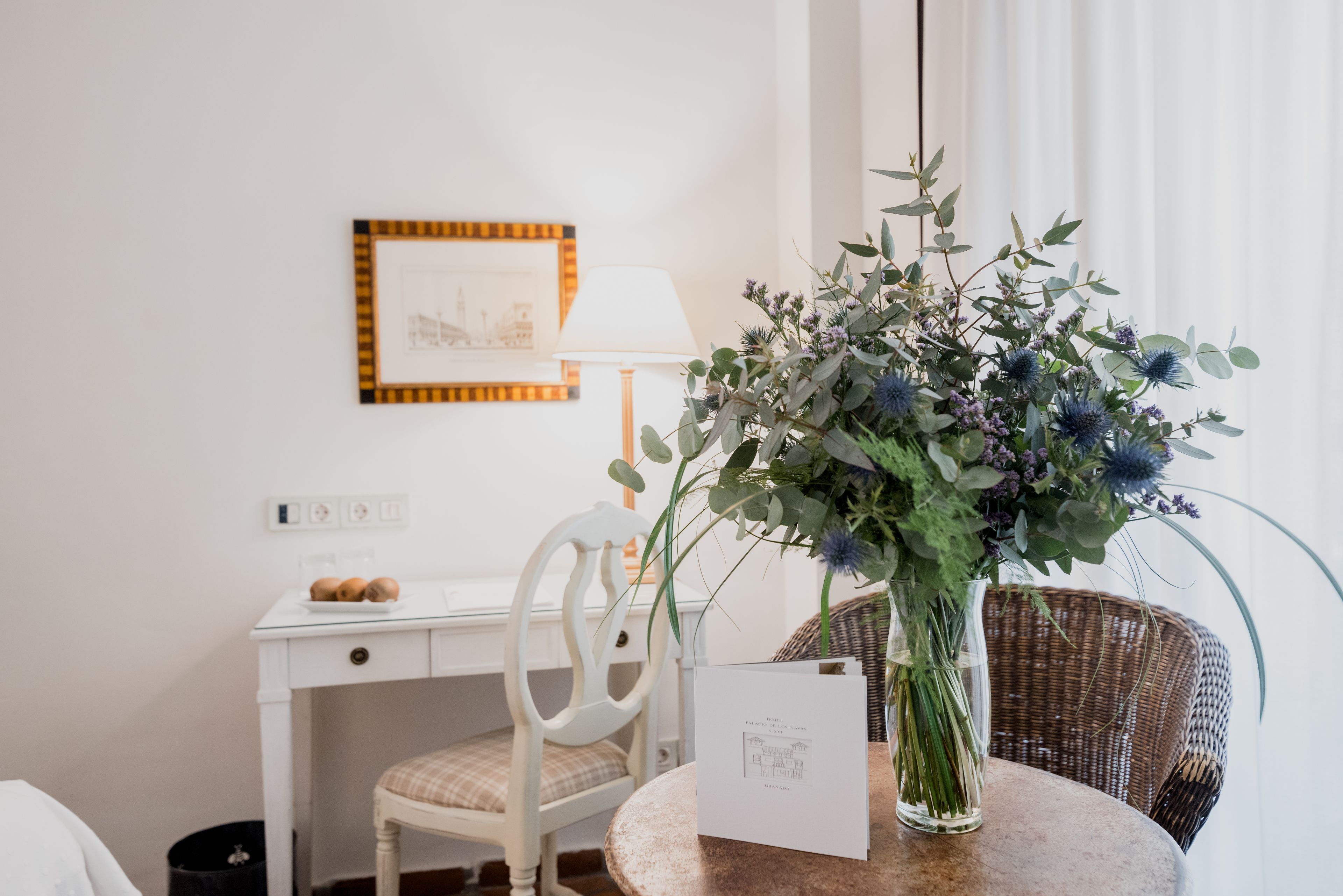 Una habitación con un escritorio blanco, una silla, una lámpara, un sillón de mimbre y un jarrón con flores.