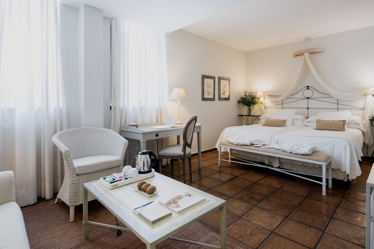 Habitación de hotel con cama, escritorio, silla, mesa con un libro y un hervidor, y ventana con cortinas.