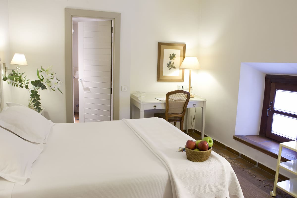 Una habitación de hotel con cama, escritorio, silla, ventana y lámpara fijada a la pared.