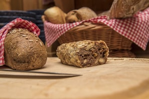 A loaf of bread and a sliced loaf on a wooden surface, with a basket of whole grains in the background. - Hotel Slalom (Bettmeralp)