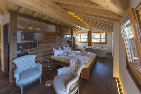 Interior de una cabaña de madera con una cama, dos sillas, una mesita, una bañera y un ventanal.