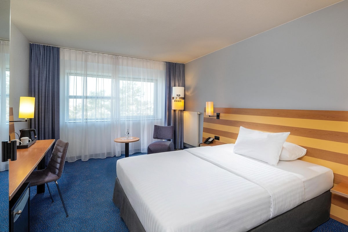 Quarto de hotel com cama, escrivaninha, cadeira, mesinha e janela com cortinas.