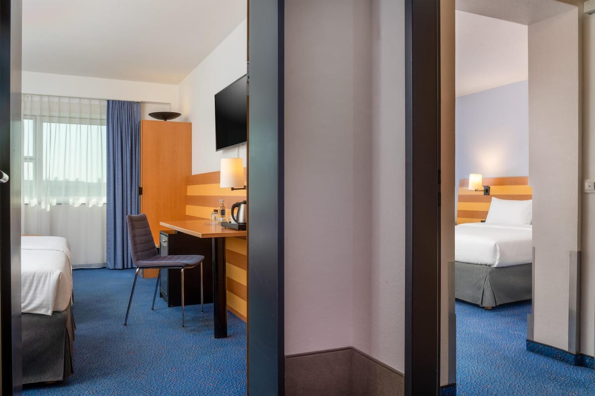Quarto de hotel com cama, escrivaninha com cadeira, televisão e janela com cortinas.