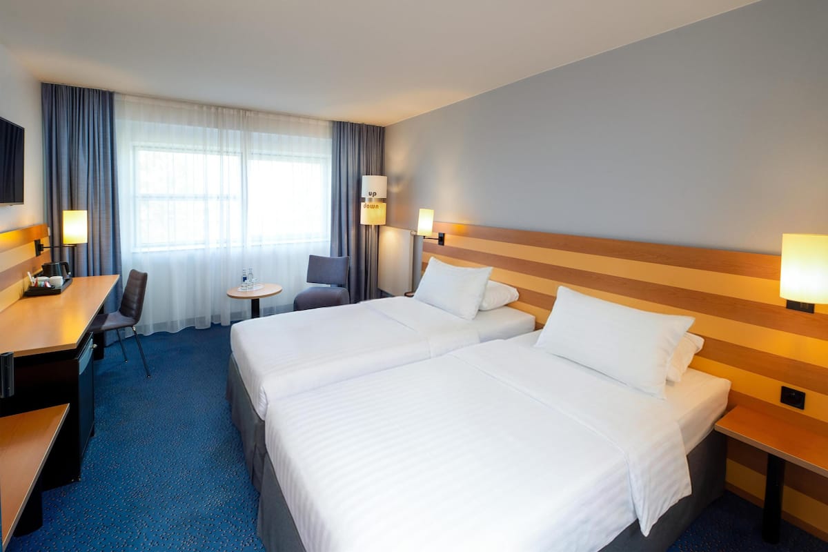 Quarto de hotel com duas camas, uma escrivaninha, uma cadeira, uma mesinha e uma televisão.