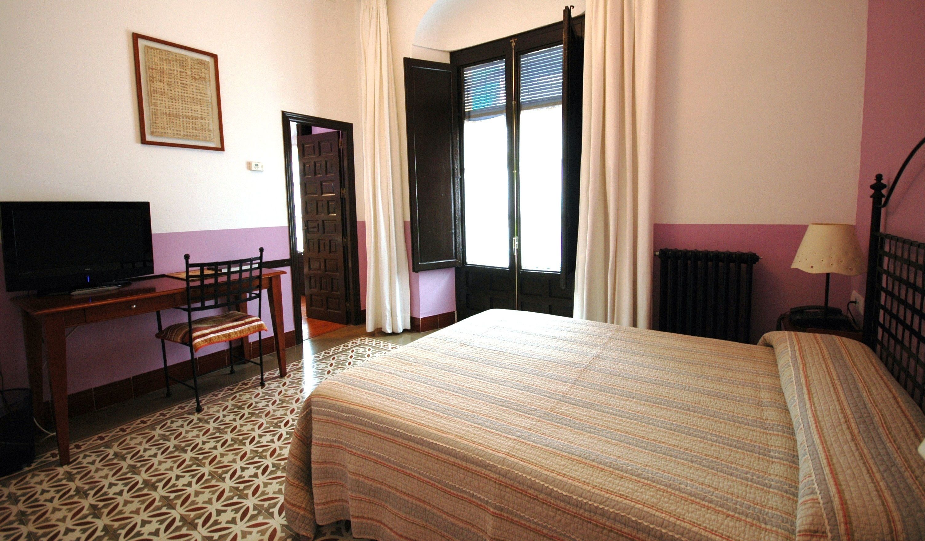 A hotel room with a bed, a desk with a chair, a television, and a window with curtains.