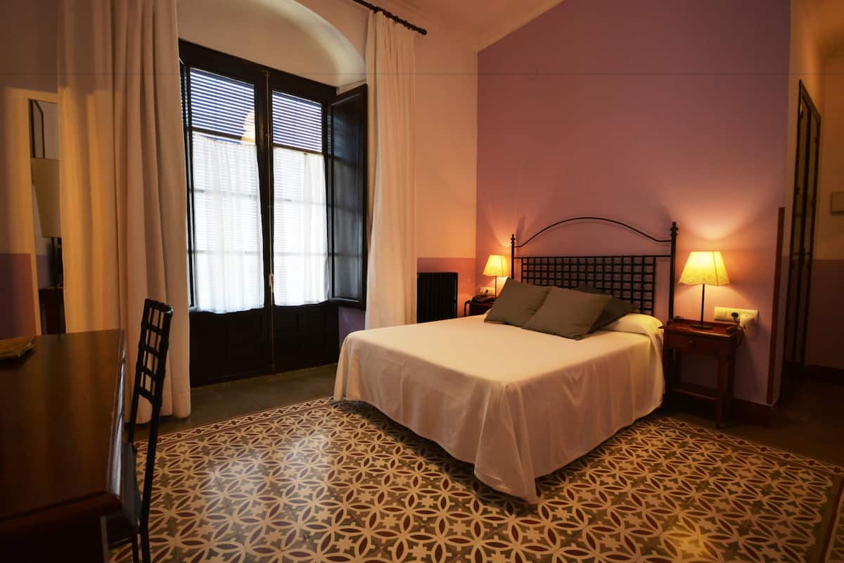 A bedroom with a bed, two bedside tables with lamps, a chair, a window with curtains, and patterned floor tiles.