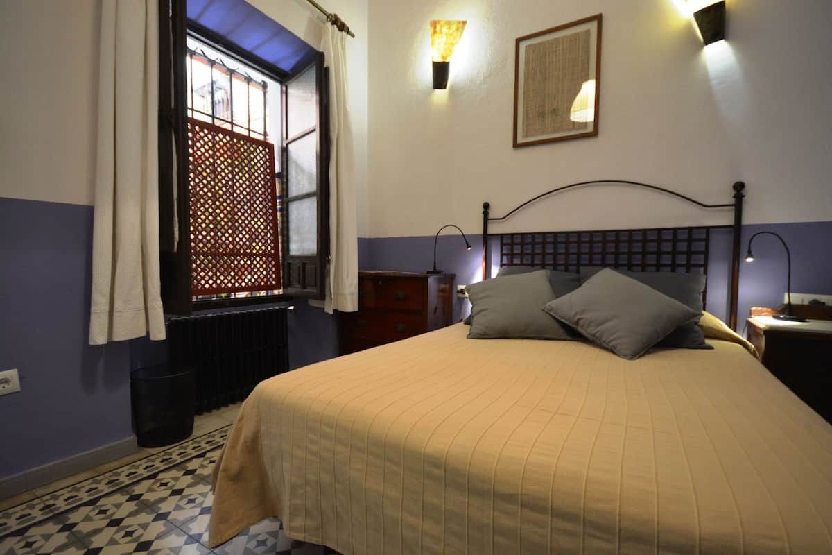 A bedroom with a bed, a window with a view, a wall-mounted light, and a framed picture.