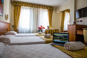 Quarto de hotel com duas camas, um laptop em uma delas, uma mesa com um vaso de flores e uma televisão na parede.