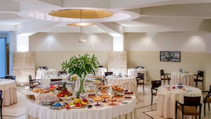 A dining area with round tables covered in white tablecloths, various food and drink items, and a central plant. - Enea Hotel Aprilia (Aprilia)