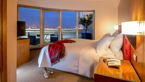 A hotel room with a large bed, a bedside table with a phone, a TV, and a view of the city at night. - Enea Hotel Aprilia (Aprilia)