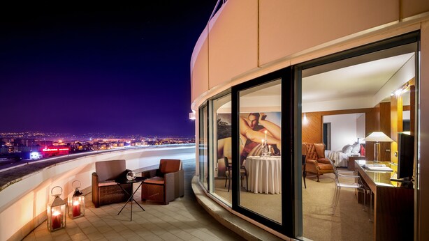 A balcony with a view of a cityscape at night, a dining area with a table and chairs, and a large window with a painting. - Enea Hotel Aprilia (Aprilia)