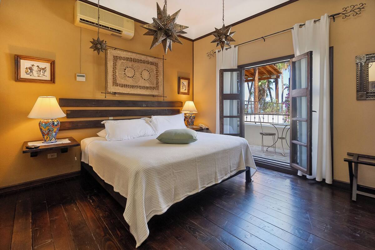 Habitación de hotel con piso de madera, cama con ropa blanca, dos lámparas de noche, una lámpara colgante con forma de estrella, una obra de arte enmarcada sobre la cama, y un balcón con vista a palmeras.