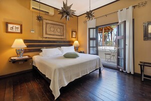 Habitación de hotel con piso de madera, cama con ropa blanca, dos lámparas de noche, una lámpara colgante con forma de estrella, una obra de arte enmarcada sobre la cama, y un balcón con vista a palmeras.