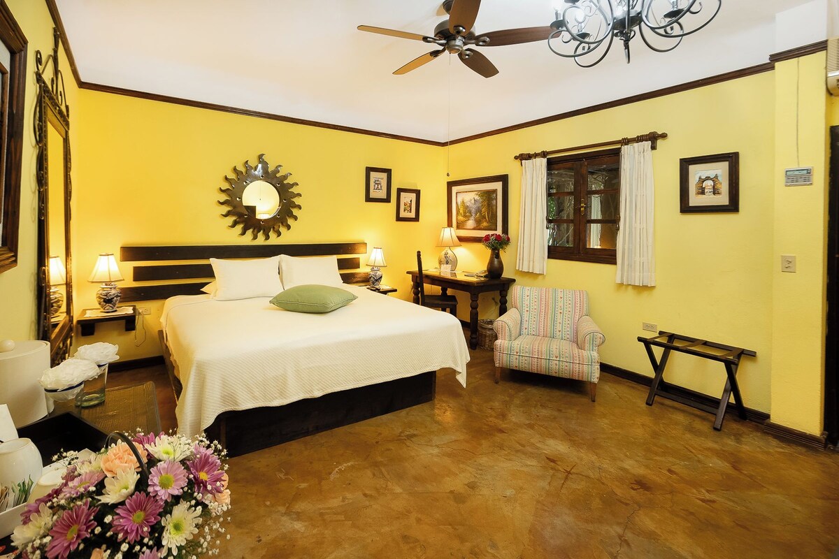 A hotel room with a bed, a chair, a table, a ceiling fan, and a window with curtains.