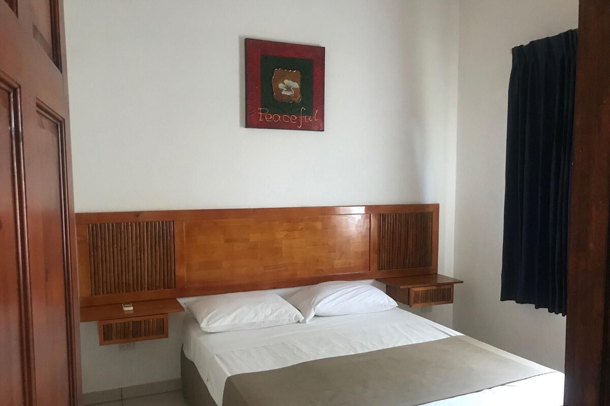 A hotel room with a wooden headboard, a bed with white linens, a nightstand, and a wall-mounted picture frame.