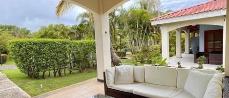 A covered outdoor seating area with a white sofa and wicker furniture, a ceiling fan, and a view of a lush garden and a building with a red roof.