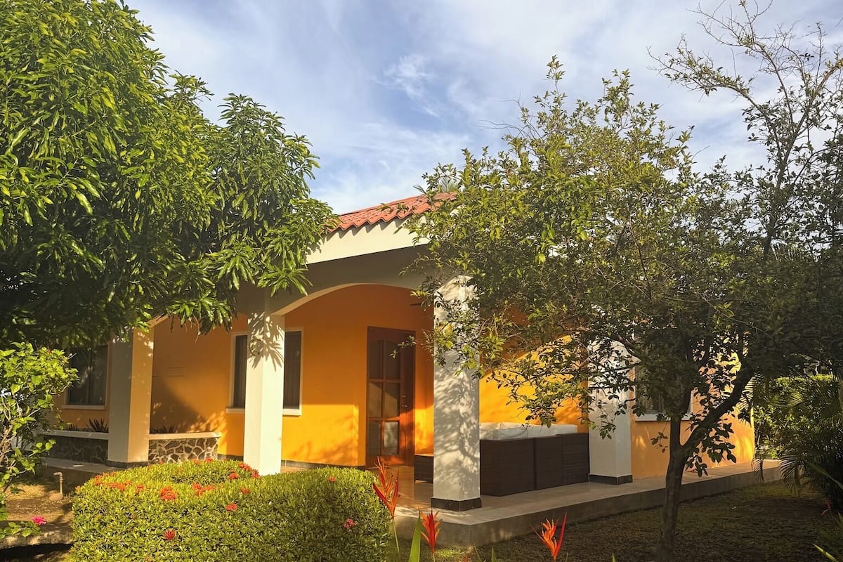 A modern building with a yellow facade, surrounded by lush greenery and flowering plants.
