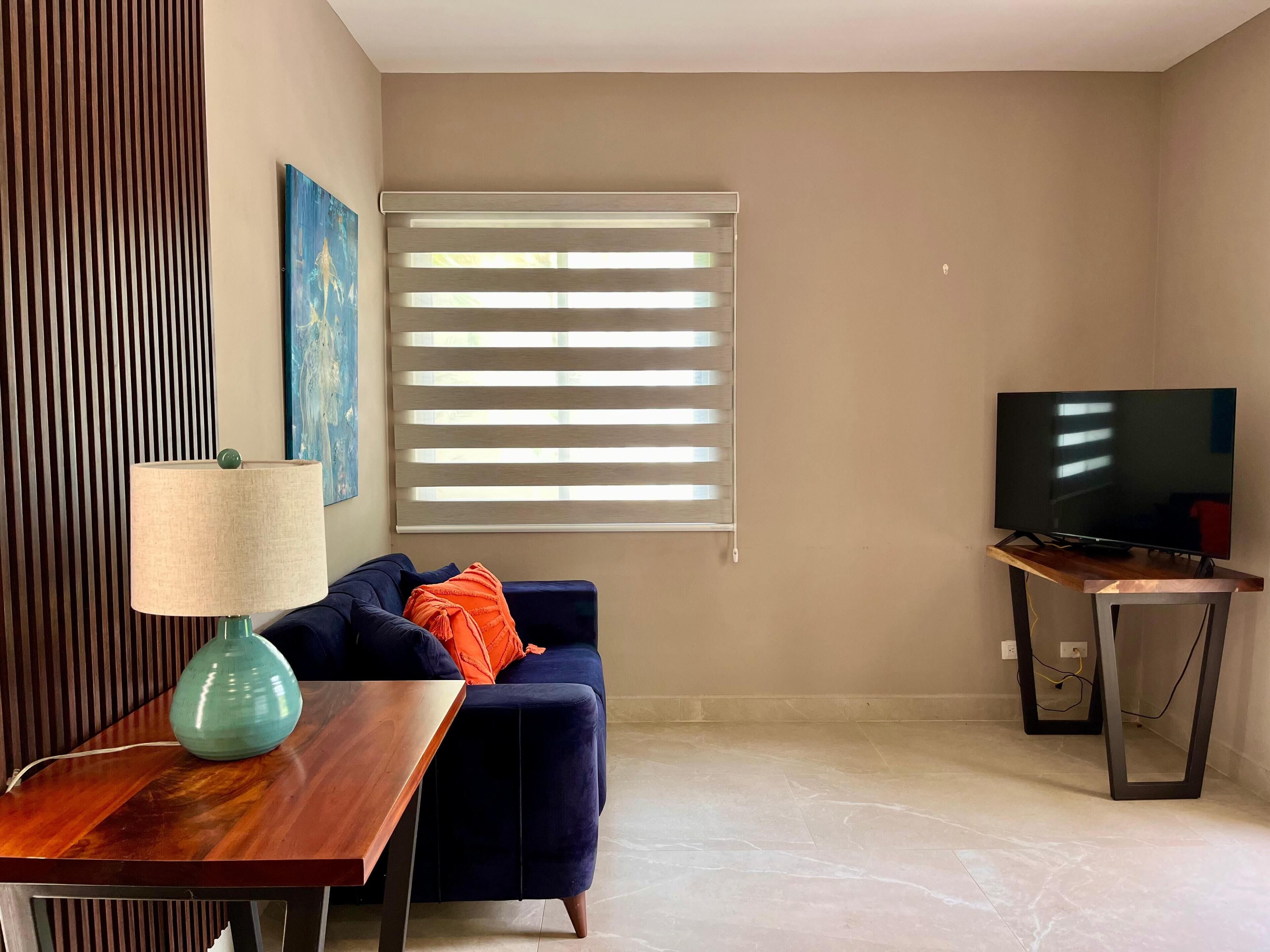 A living room with a dark blue sofa, a wooden coffee table, a teal lamp, a television on a stand, and a window with blinds.