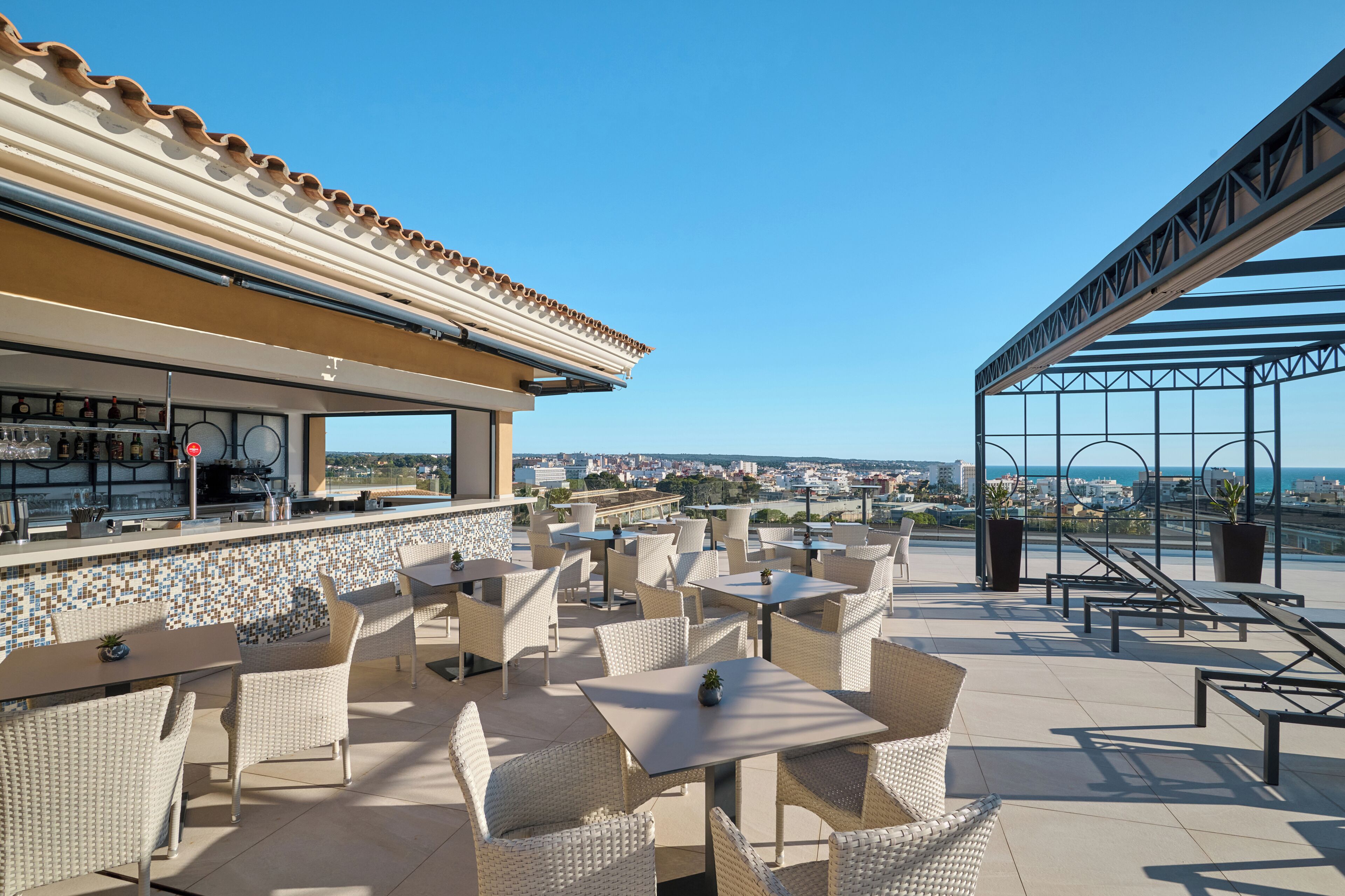 Una terraza exterior con mobiliario de mimbre, una zona de bar y vistas a la ciudad y al mar.