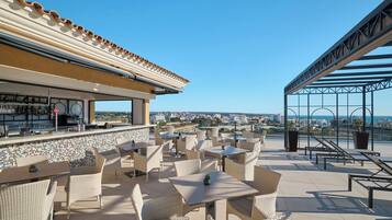 Una terraza exterior con mobiliario de mimbre, una zona de bar y vistas a la ciudad y al mar.