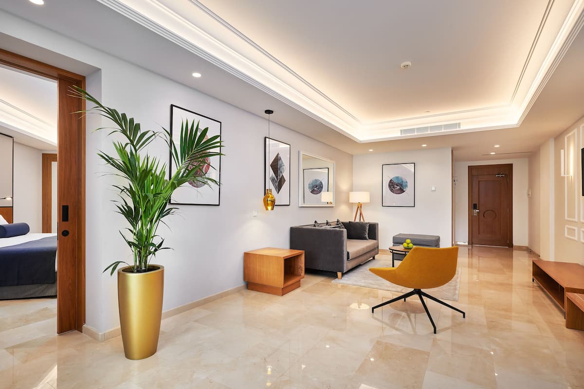 A modern hotel room with a large bed, a sitting area with a sofa and armchairs, a potted plant, and artwork on the walls.