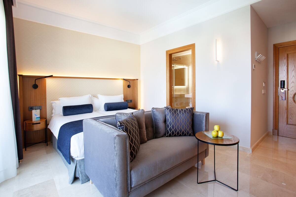 A modern hotel room with a bed, a sofa, a small table with fruit, and a wooden door.