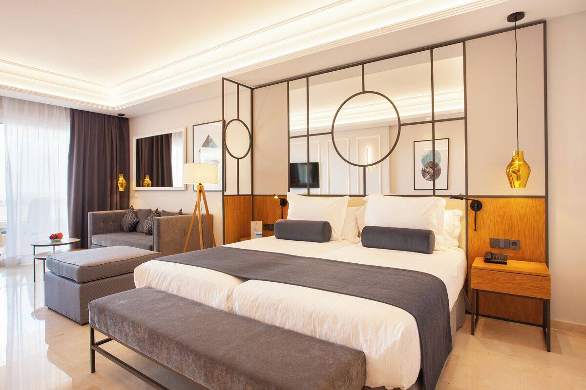 A modern hotel room with a large bed, a sofa, a bench, a bedside table, and a TV.
