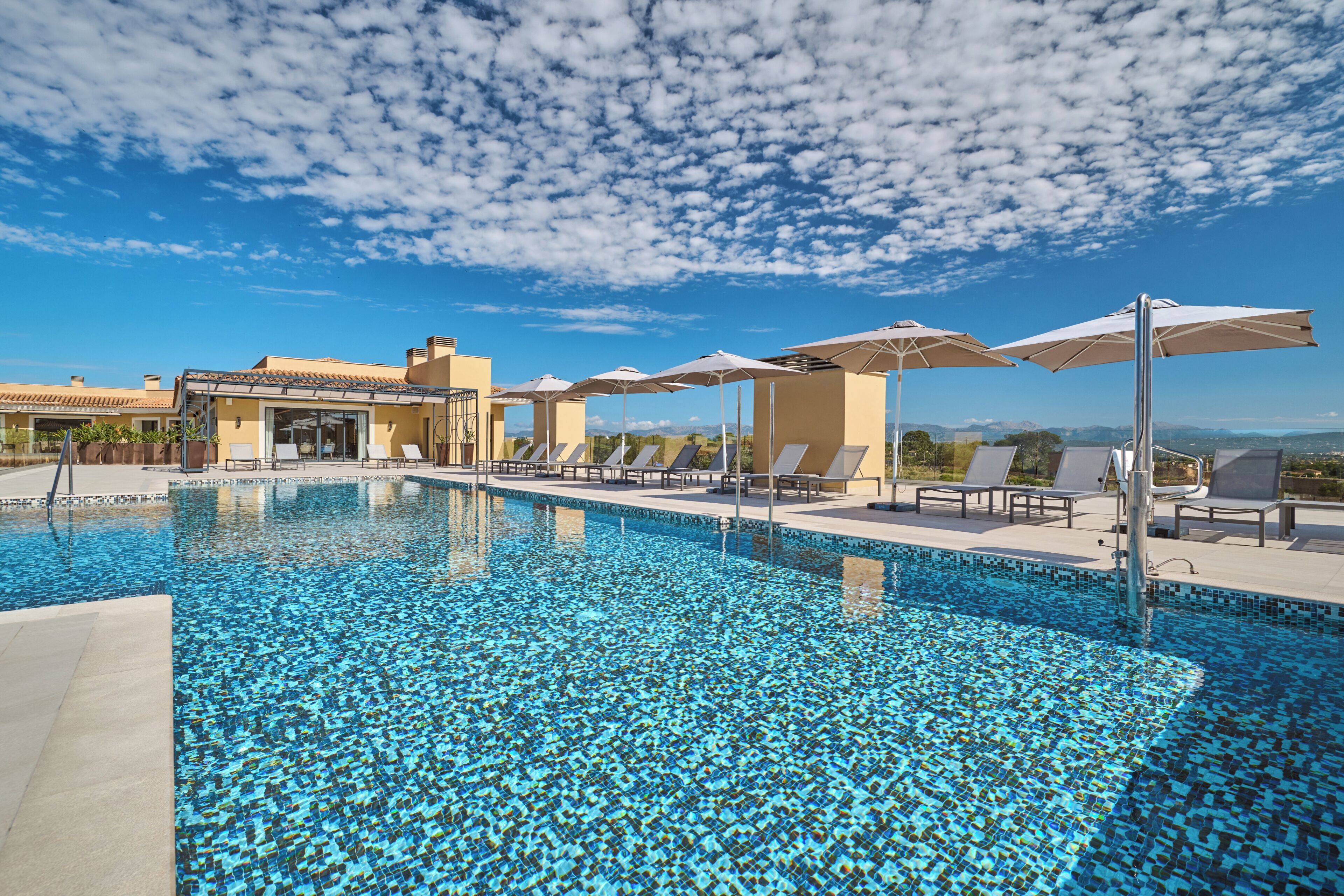 Una piscina con lettini e ombrelloni, cielo sereno e un edificio sullo sfondo.