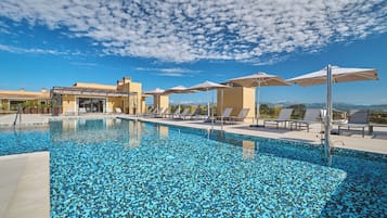 Una piscina con lettini e ombrelloni, cielo sereno e un edificio sullo sfondo.