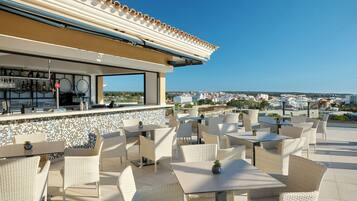 Eine Terrassengaststätte mit Korbmöbeln, einer Bar mit Fliesenrückwand und Blick auf die Stadtlandschaft.