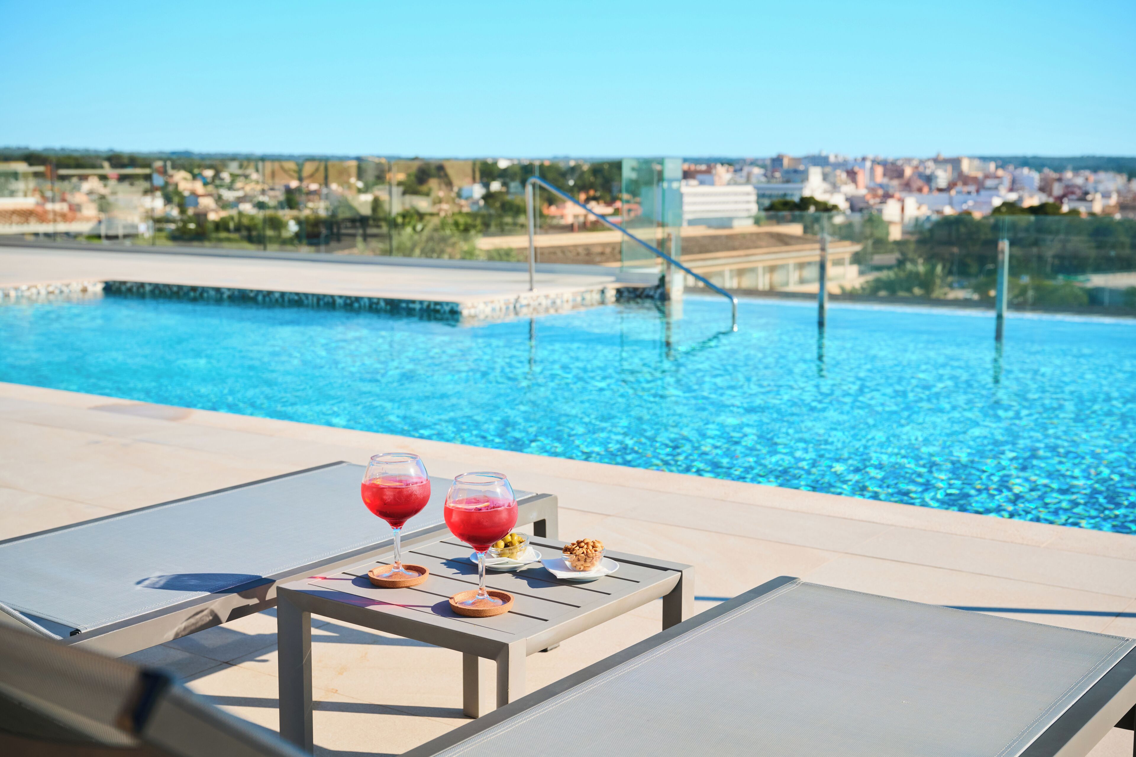 Zona relax al costat de la piscina amb dues gandules, una taula amb begudes i aperitius, i vista a la piscina i al paisatge urbà.