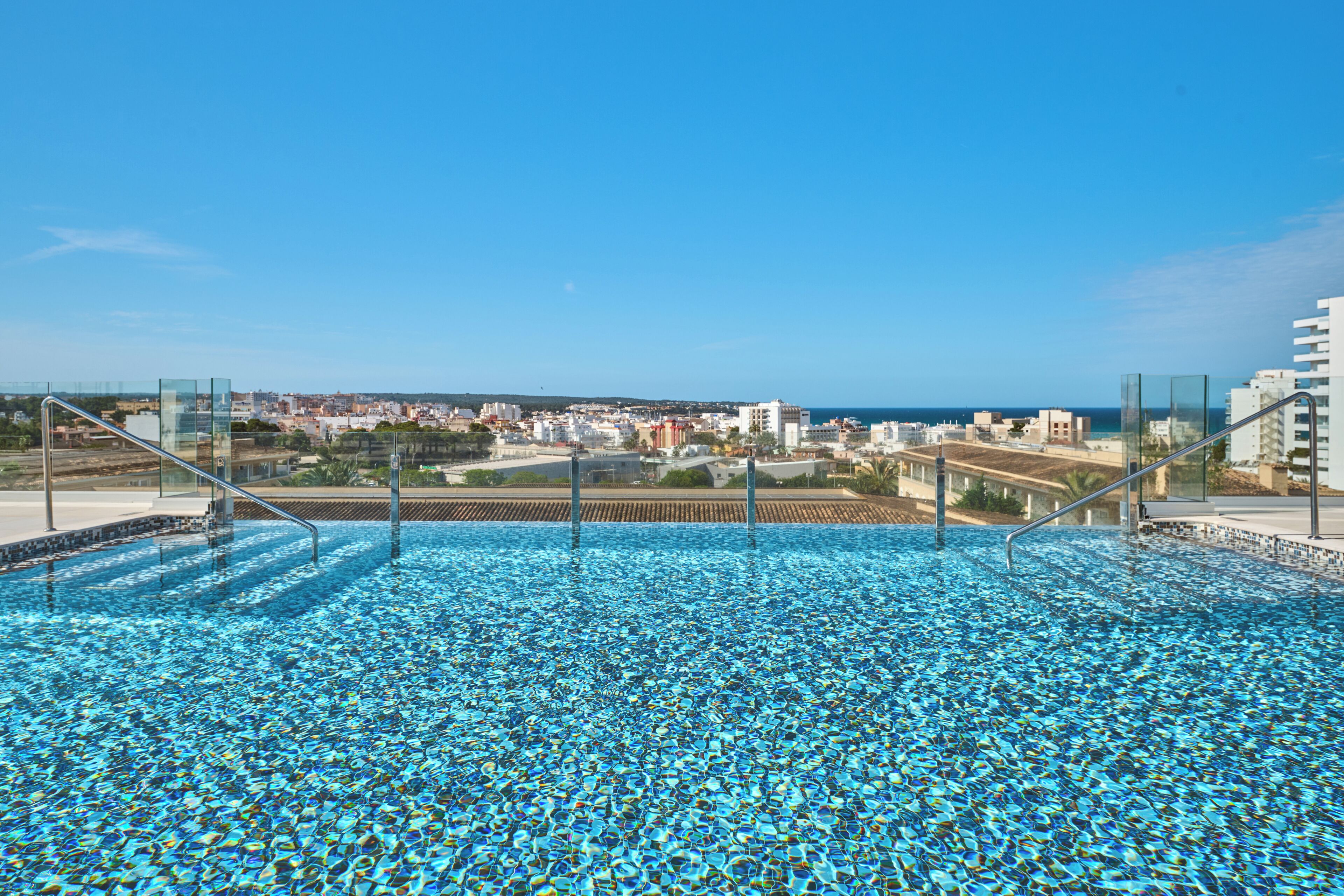 Una piscina en la azotea con agua azul cristalina, rodeada de barandillas de vidrio, con vistas a un paisaje urbano que incluye edificios y una lejana vista al mar.