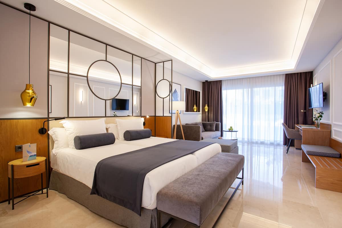 A modern hotel room with a large bed, bedside tables, a desk, a chair, and a TV.